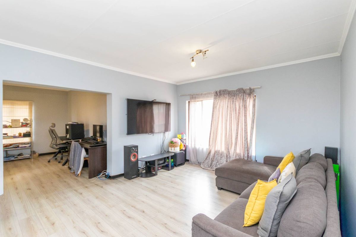 A spacious living room featuring light-toned laminate flooring, cove cornicing, and an open-plan layout connecting to an adjacent office or study area.