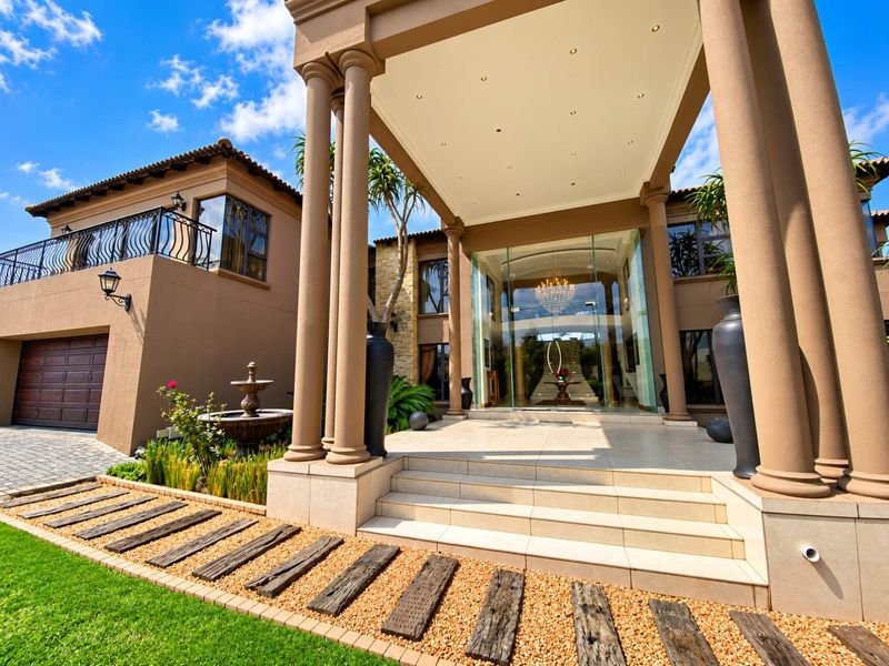 Grand front entrance featuring a covered portico with stone columns, tiled steps, and a modern glass facade, complemented by a well-maintained garden.