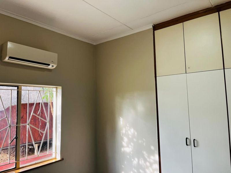 A clean, empty bedroom featuring built-in cupboards, an air conditioning unit, and a window with security bars.