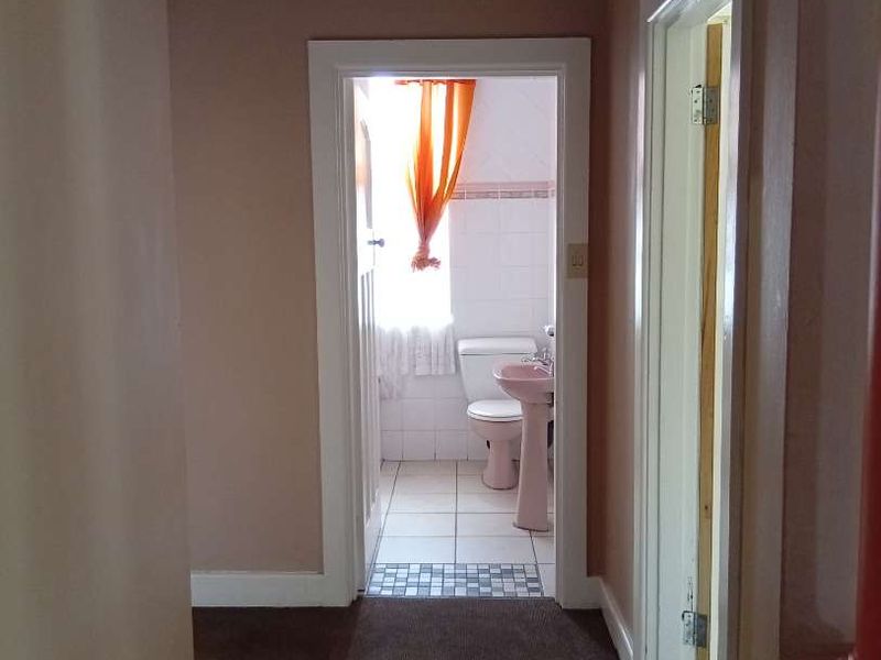 A view from a carpeted hallway looking through a doorway into a bathroom with tiled flooring and a pedestal basin.