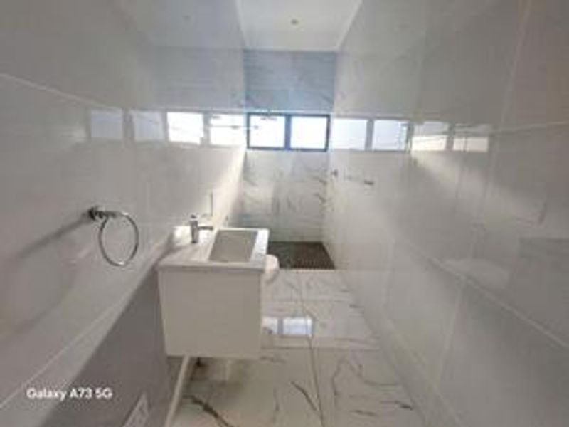 A modern, minimalist bathroom featuring white marble-look wall and floor tiles, a wall-mounted vanity with a basin, and a window for natural light.