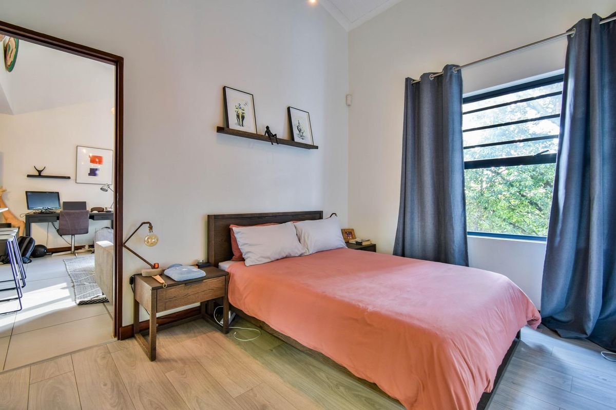 A well-lit bedroom featuring light wood-look flooring, a large window with curtains, and an adjoining room visible through the doorway.