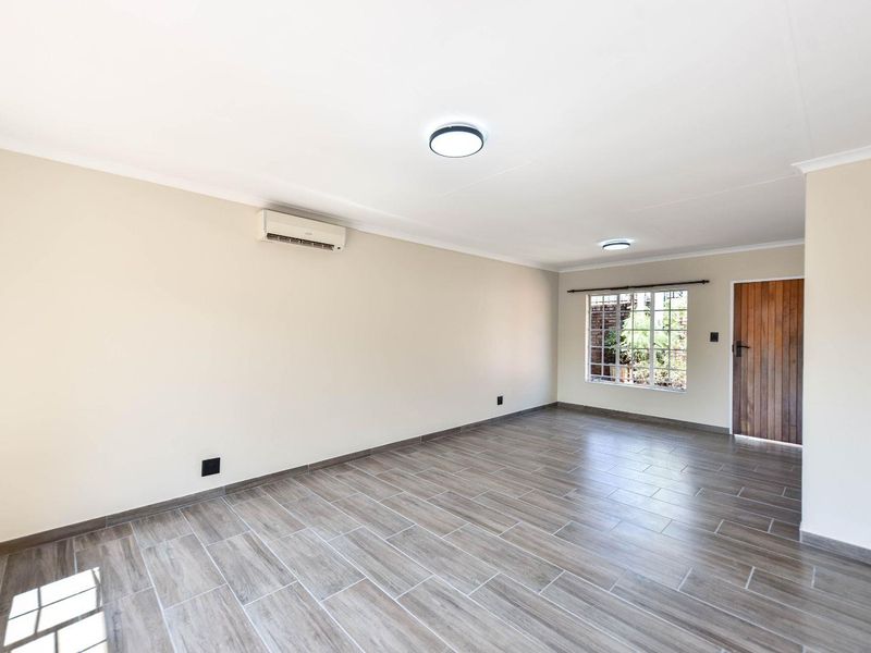 A bright, empty living room featuring modern wood-look tiled flooring, a wall-mounted air conditioning unit, and a wooden entrance door.
