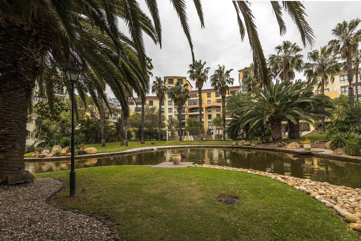 A view of the apartment complex exterior featuring a well-maintained landscaped garden with a central ornamental pond and mature palm trees.