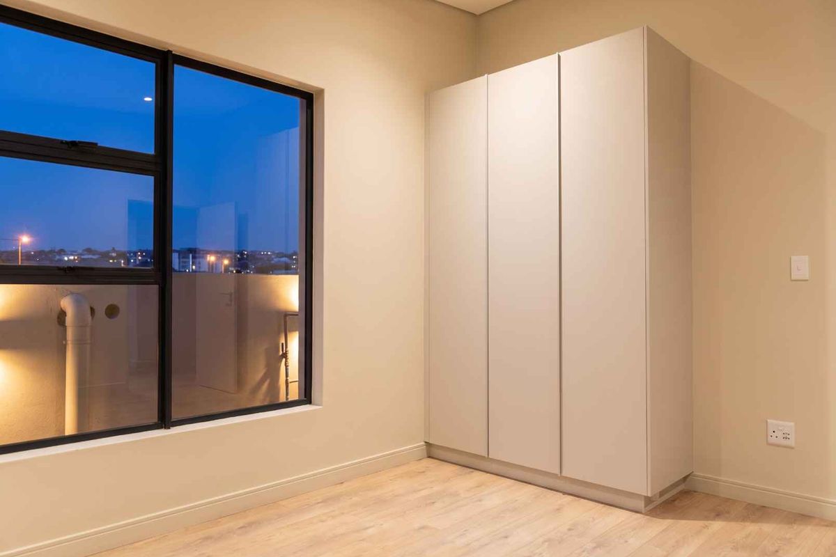 A clean, modern bedroom featuring neutral walls, light laminate flooring, and sleek built-in cupboards with large windows providing natural light.