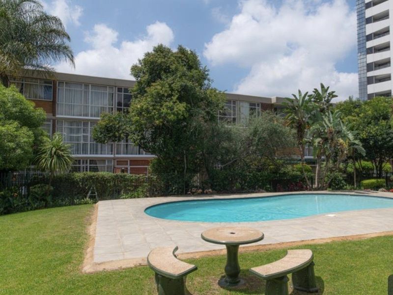 A well-maintained communal swimming pool area featuring a paved deck, surrounding lawn, and established garden landscaping within an apartment complex.