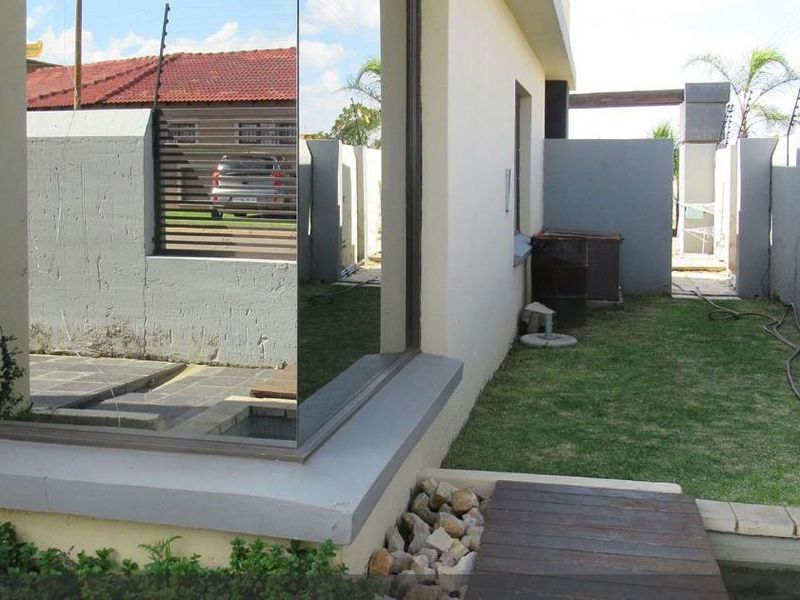 Rear exterior view of the property featuring a lawn, paved walkway, boundary wall with electric fencing, and a large reflective glass window.