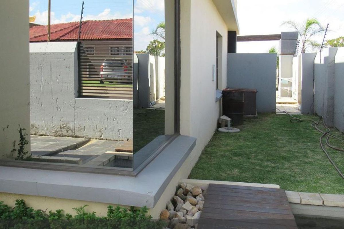Rear exterior view of the property featuring a lawn, paved walkway, boundary wall with electric fencing, and a large reflective glass window.