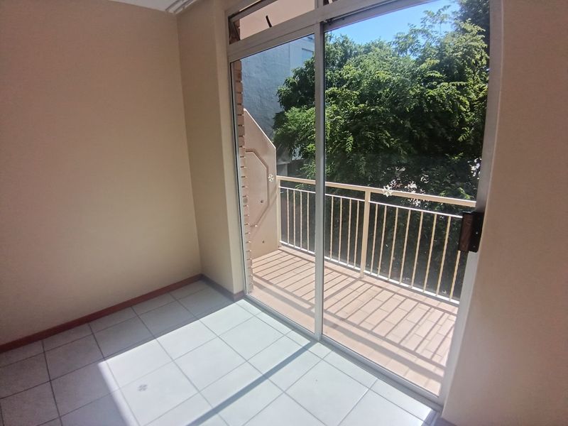 A bright, empty balcony area featuring tiled flooring, a sliding glass door, and a metal balustrade overlooking greenery.