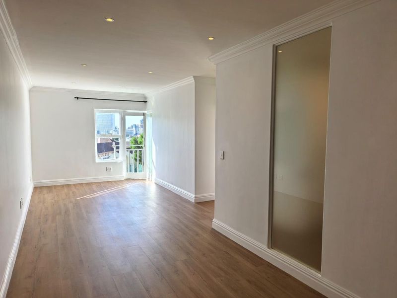 A bright, empty living area featuring modern laminate flooring, recessed downlighting, decorative cornicing, and a glass-paneled door leading to a balcony.