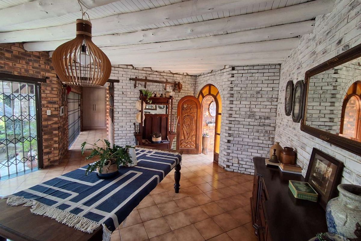 A rustic living area featuring exposed timber beam ceilings, painted brick walls, and ceramic tile flooring with an arched wooden entrance door.