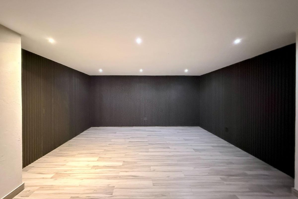 A spacious, empty room featuring modern dark vertical slat wall paneling and light-toned wood-look flooring, illuminated by recessed ceiling spotlights.