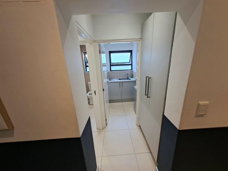 A clean hallway leading to a small kitchenette area and bathroom, featuring tiled floors and built-in storage cabinetry.