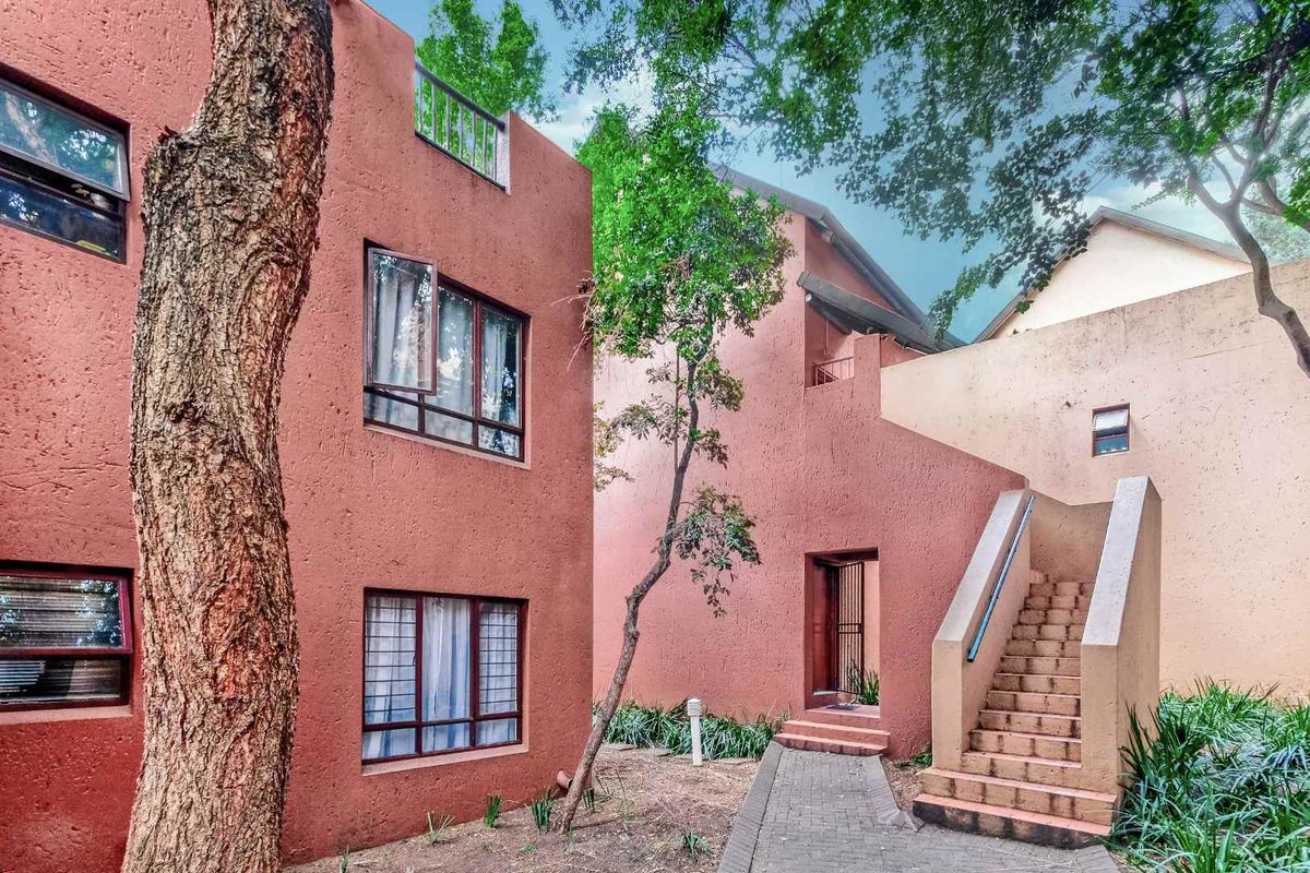 Exterior view of a multi-story apartment complex featuring terracotta-toned textured walls, paved walkways, and external staircases surrounded by mature trees.