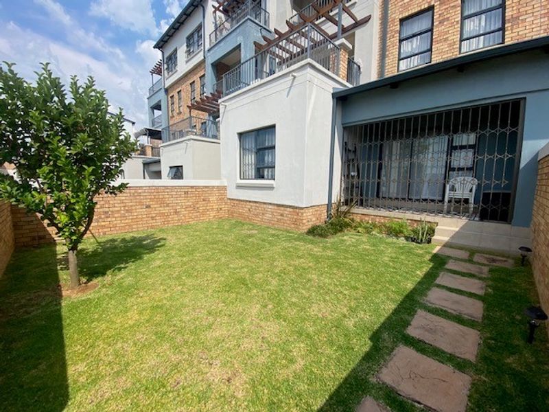 A well-maintained rear garden featuring a lawn, brick boundary wall, paved stepping stones, and a covered patio area with security gate access.