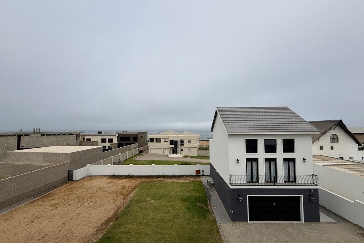 Aerial view of a modern two-story residential property featuring a paved driveway, garage, landscaped lawn, and a balcony overlooking the coastal neighborhood.