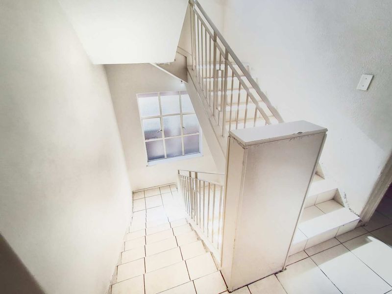 A bright, empty stairwell featuring white tiled flooring, a metal balustrade, and a grid-style window providing natural light.