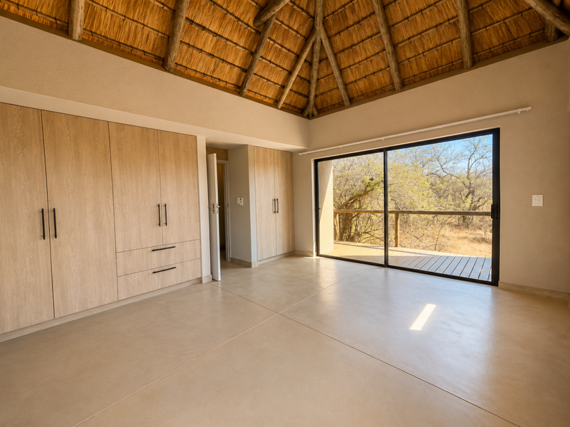 Spacious bedroom featuring a high thatched ceiling with exposed rafters, built-in wooden cupboards, polished concrete floors, and large sliding doors opening to a deck.