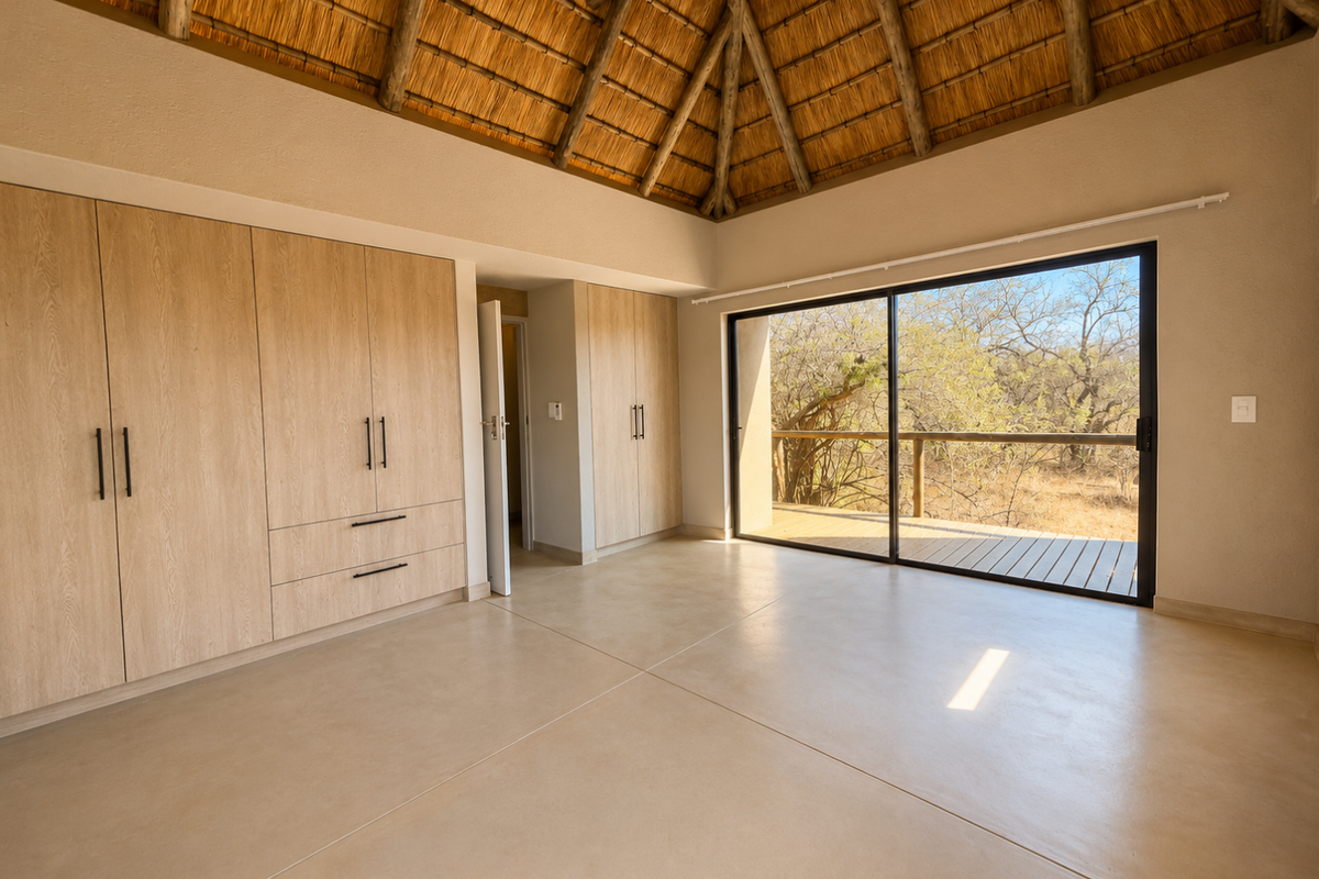 Spacious bedroom featuring a high thatched ceiling with exposed rafters, built-in wooden cupboards, polished concrete floors, and large sliding doors opening to a deck.