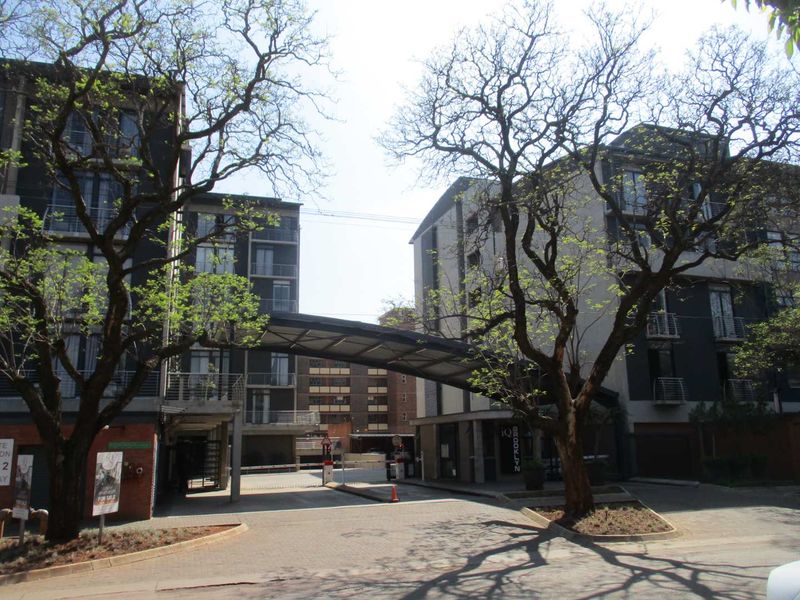 Street view of the apartment complex entrance featuring a paved driveway, security gate, and modern multi-story building facades with balconies.