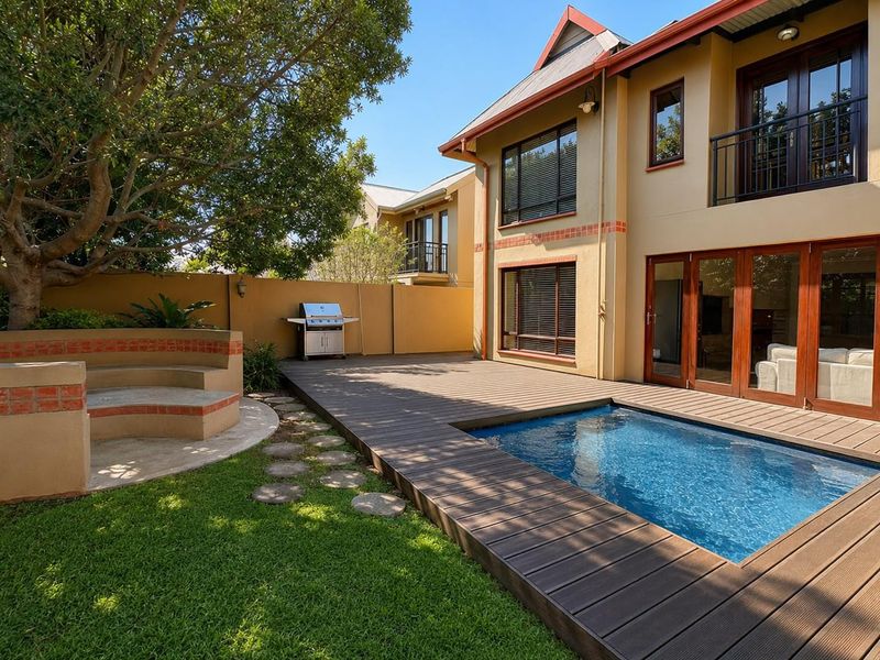 A well-maintained rear garden featuring a swimming pool, timber decking, built-in brick seating, and a double-storey house facade with balcony access.