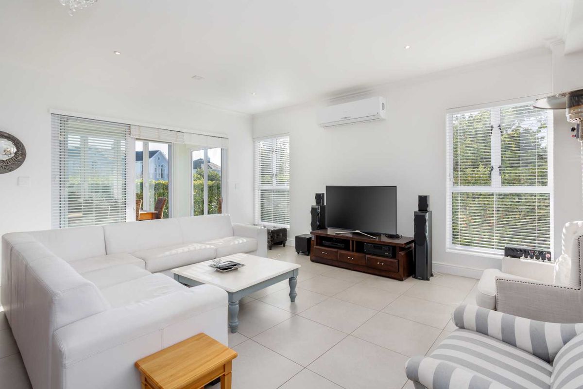 A bright and spacious living room featuring tiled flooring, recessed downlighting, a wall-mounted air conditioning unit, and large windows with venetian blinds.