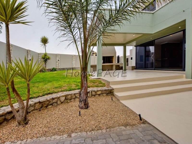 Rear exterior view featuring a covered patio with a built-in braai, stone retaining wall, landscaped garden, and sliding glass doors leading to the interior.