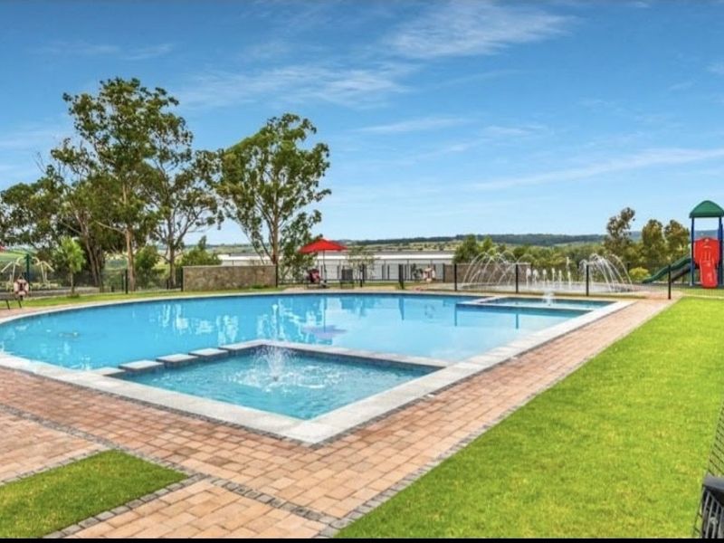 A well-maintained communal swimming pool area featuring paved surrounds, water features, and landscaped lawn, set within a secure fenced environment.