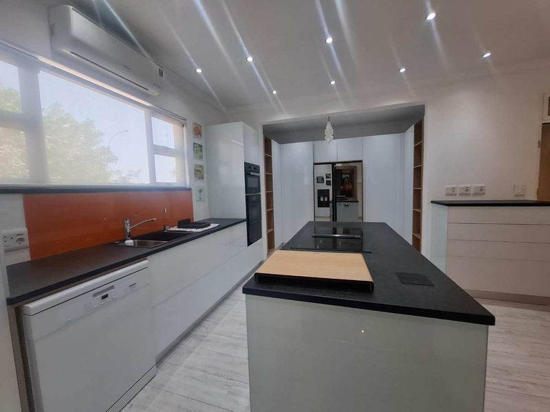 Modern kitchen featuring a central island with hob, sleek white cabinetry, dark countertops, orange splashback, and integrated appliances in a bright, open-plan layout.