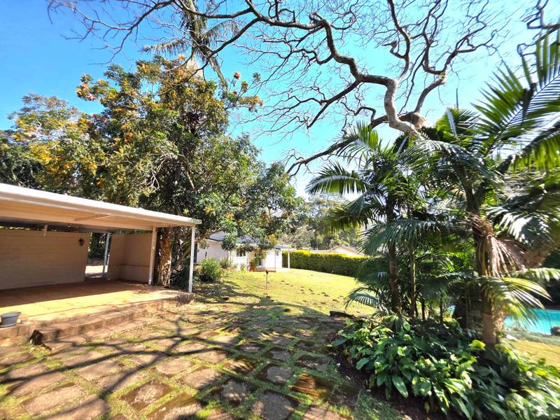 A spacious rear garden featuring a covered patio area, stone paving, and mature trees, offering a tranquil outdoor living space.