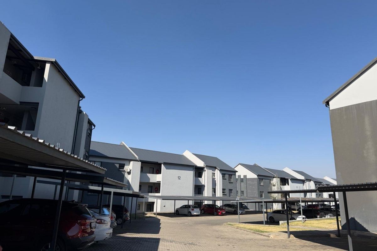 Exterior view of a multi-story apartment complex featuring paved driveways and covered parking bays under a clear blue sky.