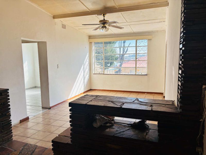 An empty living room featuring tiled flooring, a stone fireplace, and a ceiling fan, with large windows providing natural light.