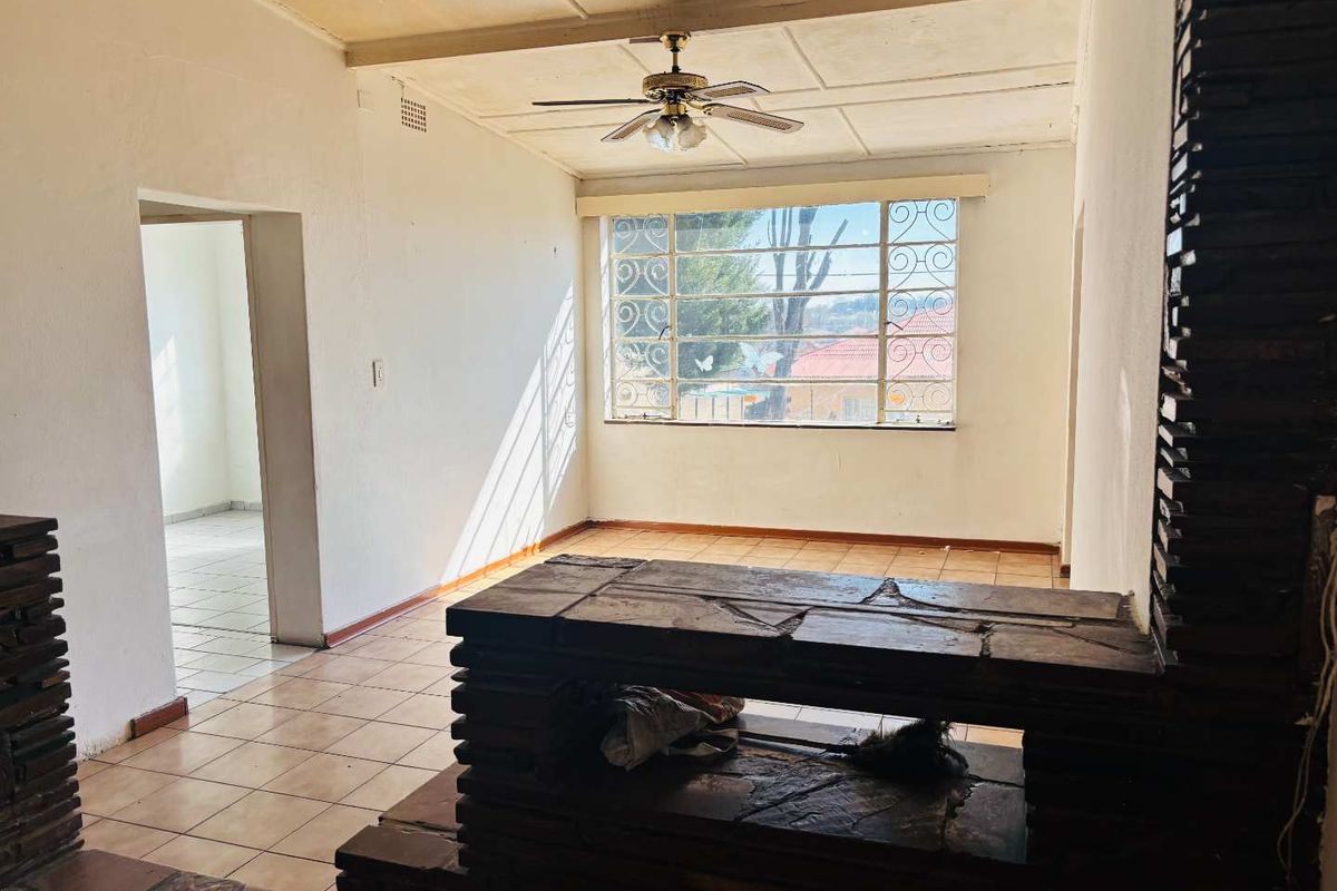 An empty living room featuring tiled flooring, a stone fireplace, and a ceiling fan, with large windows providing natural light.