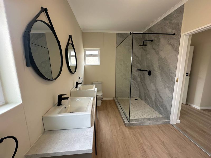 Modern bathroom featuring a double vanity with black tapware, a spacious walk-in shower with grey marble-look tiling, and light wood-look flooring.
