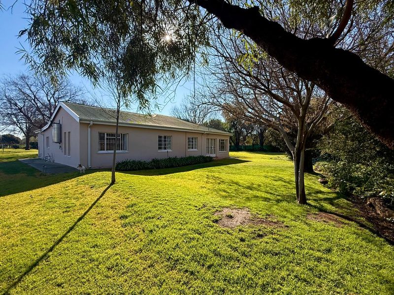 Rear view of the farm residence featuring a metal roof, security-fitted windows, and a spacious surrounding lawn area with mature trees.