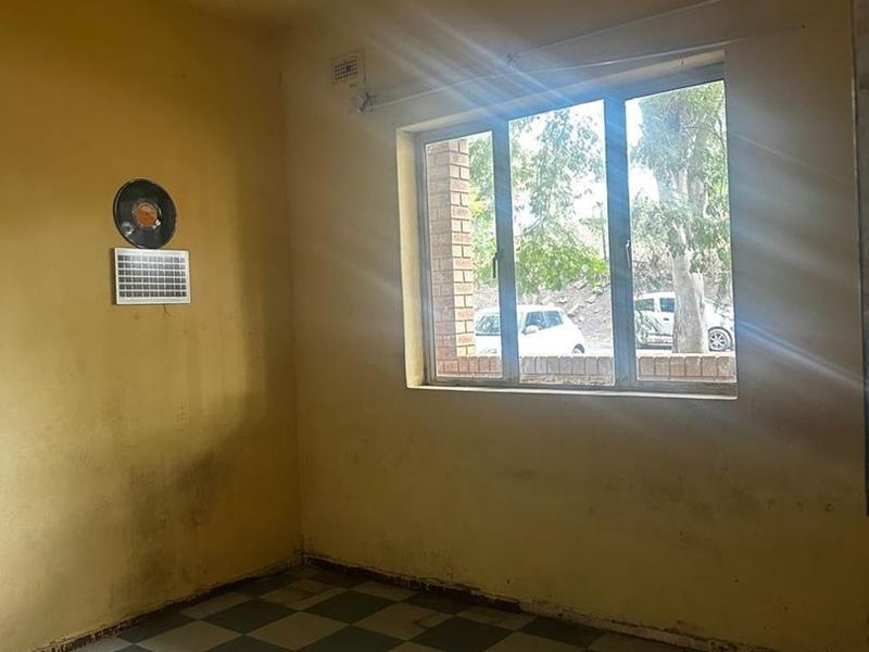 An empty living room featuring checkered tiled flooring and a window, currently requiring significant cosmetic updates and wall repairs.