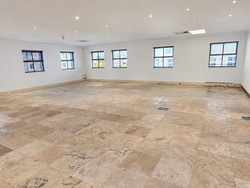 A spacious, empty office area featuring natural stone tile flooring, recessed ceiling lighting, and multiple windows providing ample natural light.