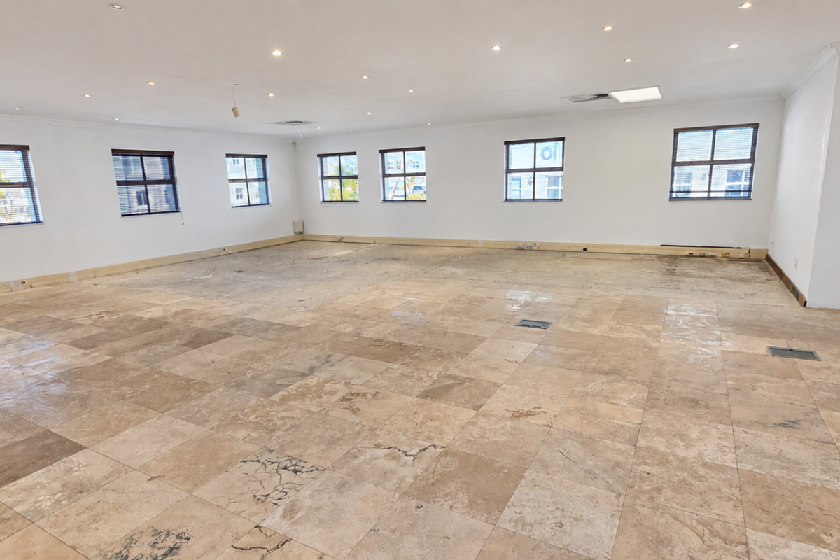 A spacious, empty office area featuring natural stone tile flooring, recessed ceiling lighting, and multiple windows providing ample natural light.