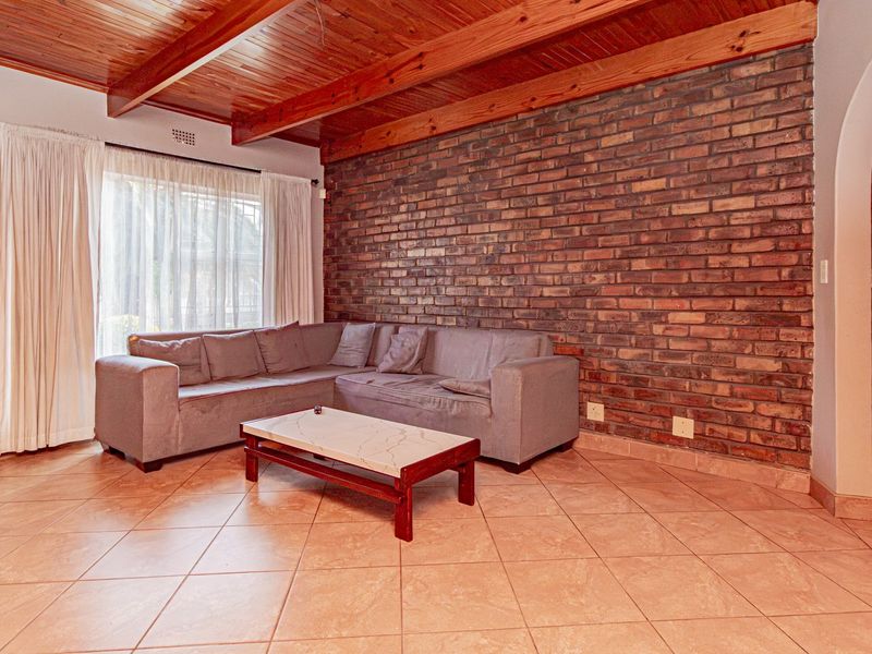 Spacious living room featuring a rustic exposed brick wall, warm timber beam ceiling, and neutral ceramic tile flooring with an elegant arched entryway.