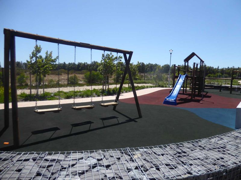 A well-maintained residential playground area featuring rubberized safety surfacing, swing sets, a slide, and decorative gabion stone walling.