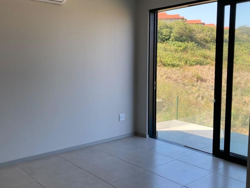 A bright, empty living area featuring neutral tiled flooring, a wall-mounted air conditioner, and large sliding glass doors opening to an outdoor space.