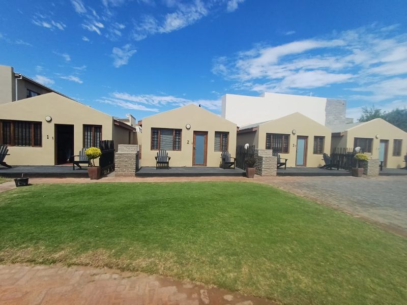 A row of single-story units featuring timber decking, brick pillars, and wooden window frames, set against a well-maintained lawn and paved driveway.