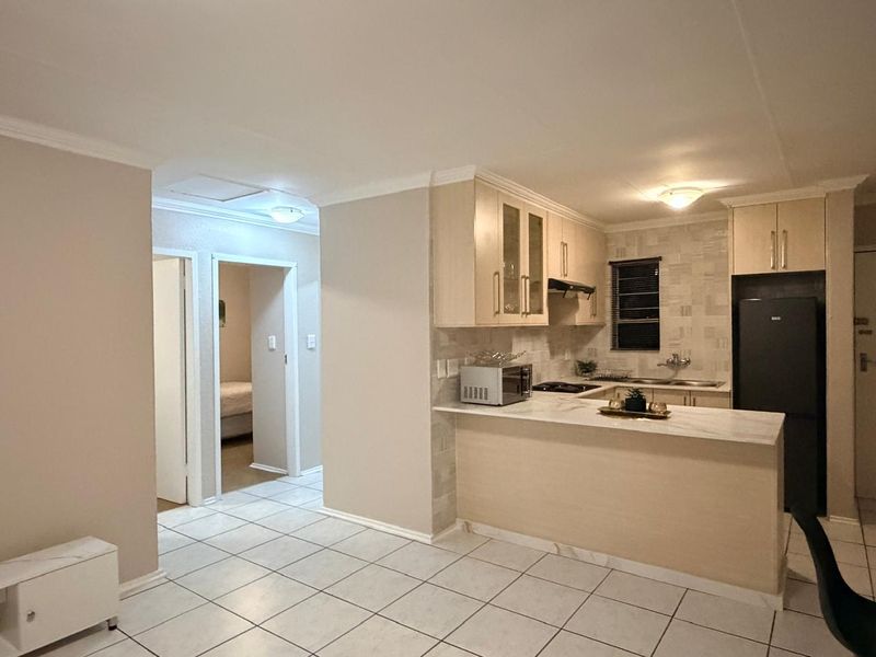 Open-plan kitchen featuring light-toned cabinetry, stone-look countertops, a breakfast bar, and ceramic floor tiles, seamlessly connecting to the hallway and adjacent living space.