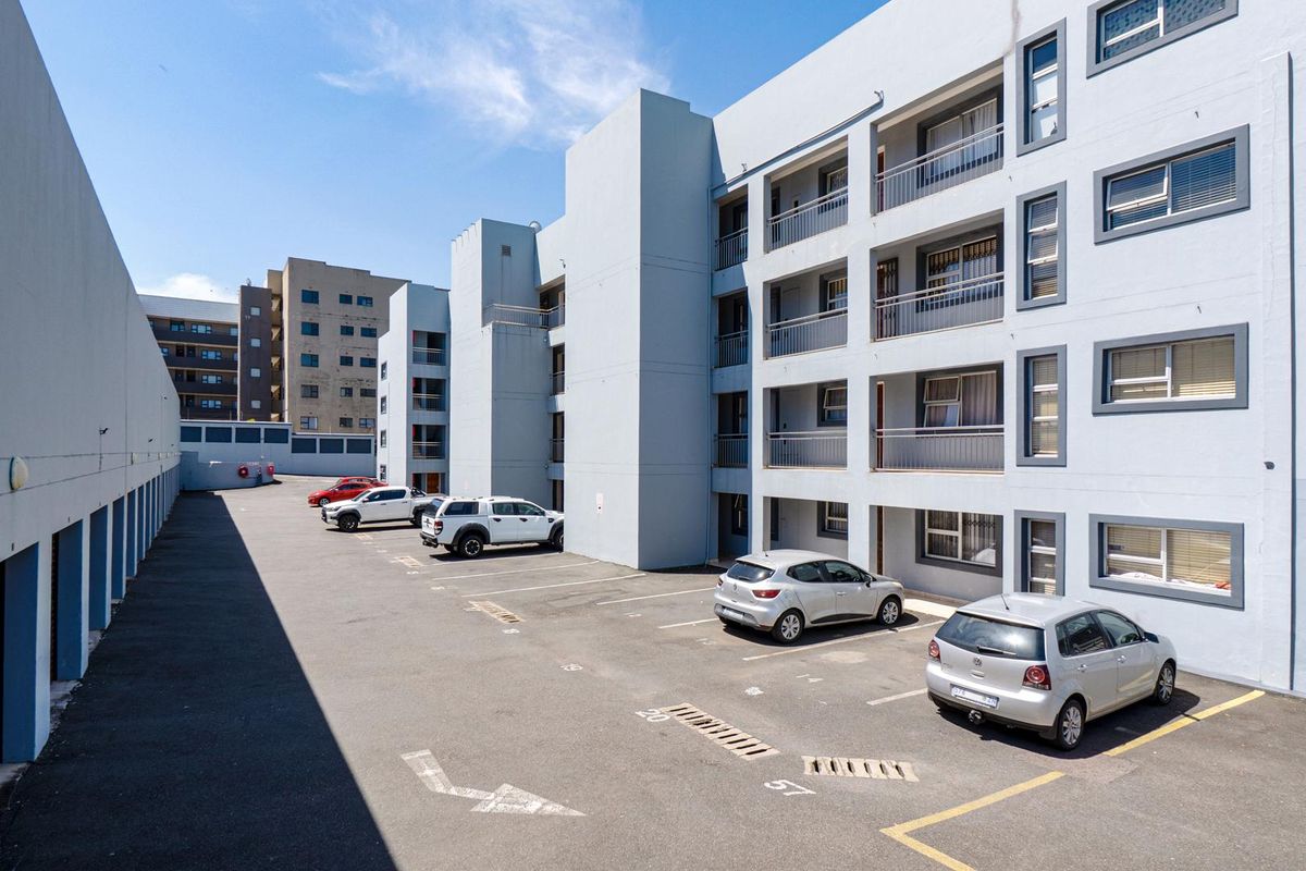 Exterior view of the apartment complex featuring a paved parking area, multi-level building facade with balconies, and designated parking bays.
