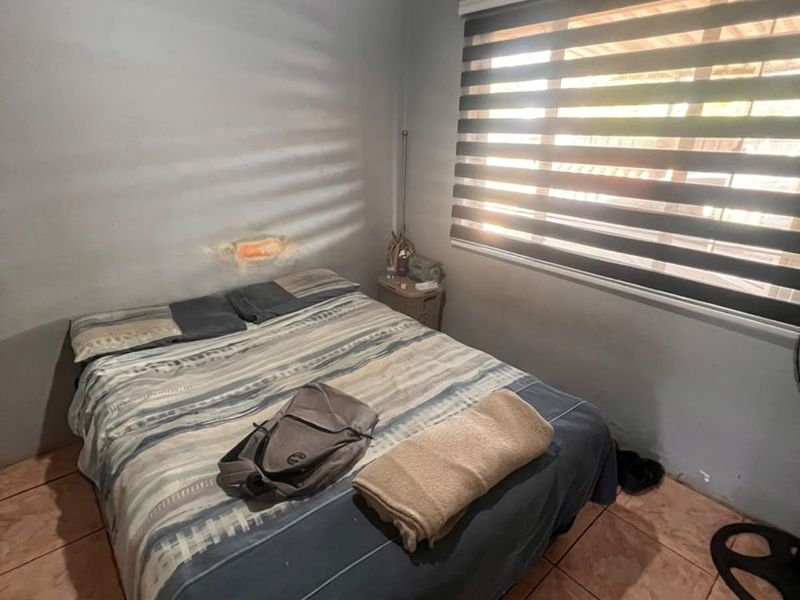 A bedroom featuring tiled flooring, a ceiling fan, and zebra blinds, currently showing visible wall damage above the bed area.