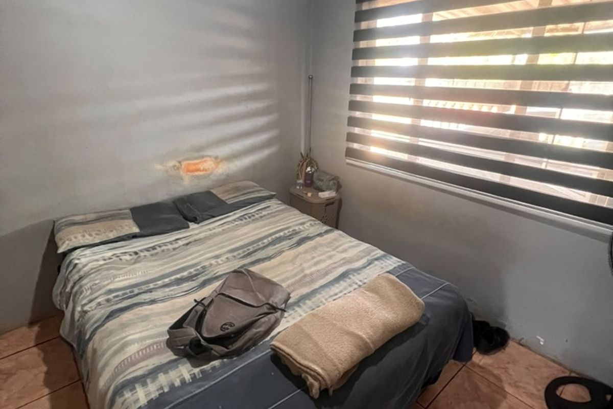 A bedroom featuring tiled flooring, a ceiling fan, and zebra blinds, currently showing visible wall damage above the bed area.