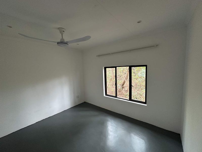 A clean, empty bedroom featuring a ceiling fan, recessed lighting, and a window with a dark frame, finished with painted concrete flooring.