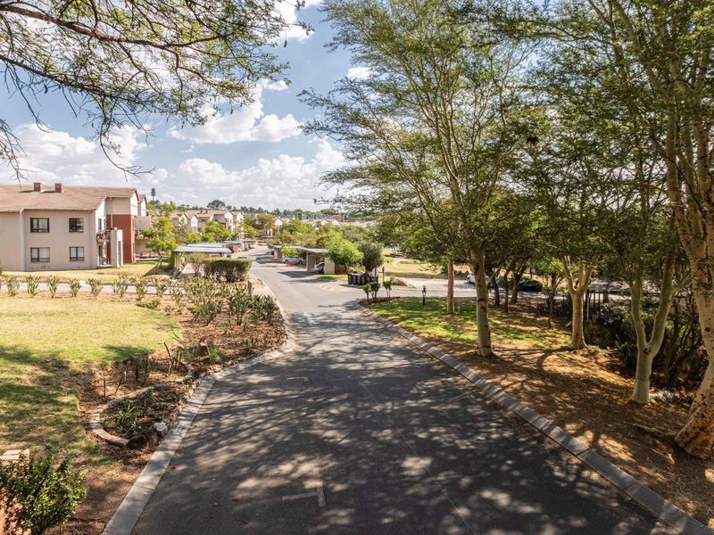 An elevated view of the apartment complex grounds, featuring a paved driveway, mature trees, landscaped garden areas, and covered parking facilities.