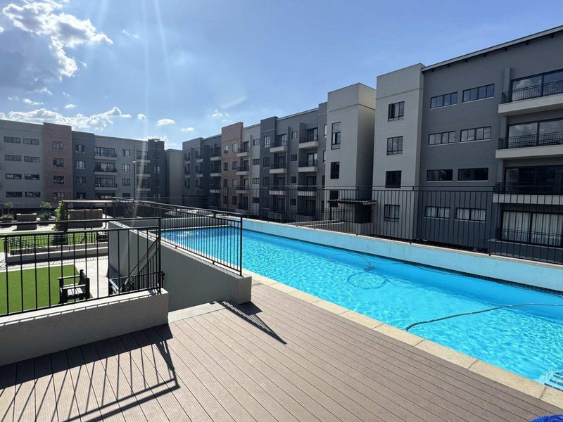 A well-maintained communal swimming pool area featuring composite decking, metal safety railings, and artificial lawn, set within a modern apartment complex.