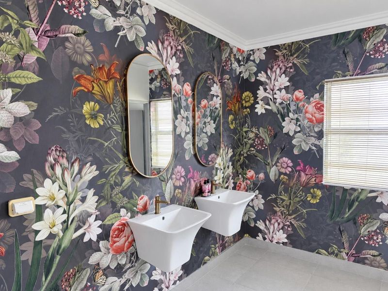A modern bathroom featuring dual wall-mounted basins with brass fixtures, set against a bold floral wallpaper and complemented by neutral floor tiles.